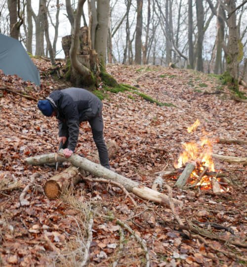 Deelnemers zagen hout bij kampvuur met tenten op achtergrond tijdens MTA 3-daagse Eifel